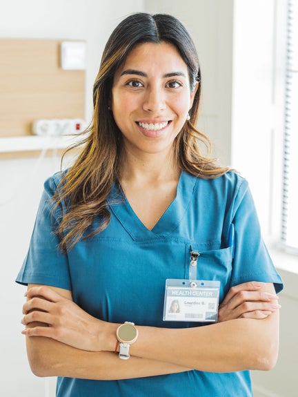 A woman in a blue scrub suit stands confidently in front of a hospital, ready to assist patients and provide care.