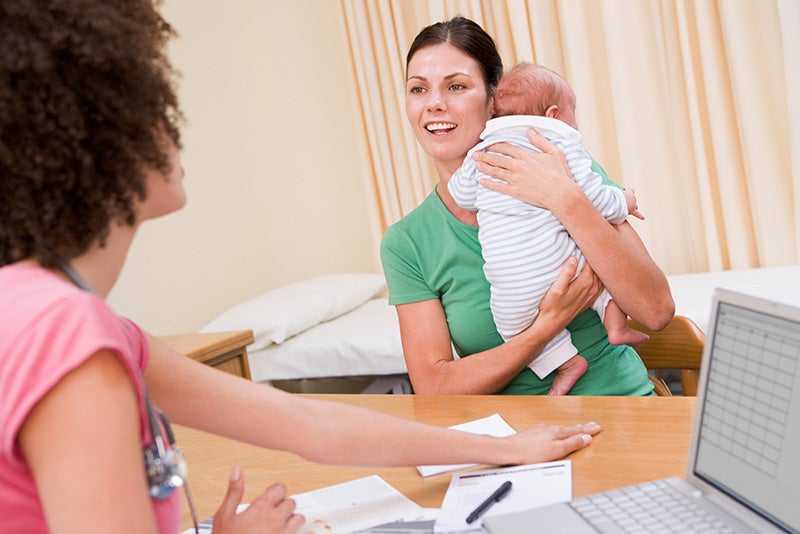 A woman sitting at a desk, gently holding a baby in her arms, smiling as she works.
