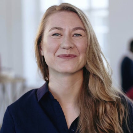 A woman with long hair smiles warmly while seated at her desk in a modern office environment.