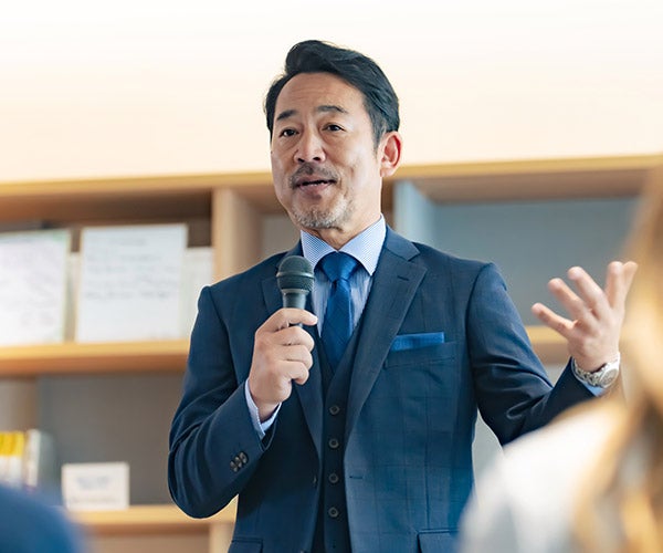 A man in a suit speaks into a microphone during a formal event, engaging the audience with his presentation.