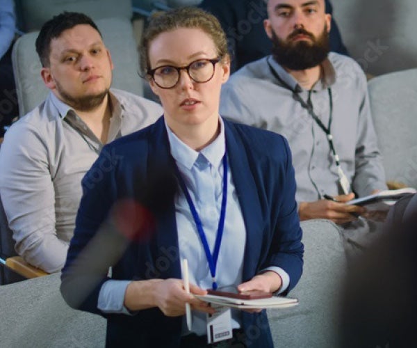A woman in glasses and a suit addresses a group of attentive listeners during a presentation.
