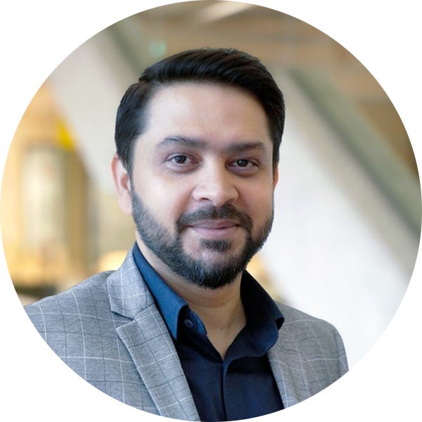 A man with a beard wearing a suit jacket, standing confidently with a neutral expression.