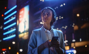 A woman stands in a city at night, illuminated by streetlights, holding her phone and looking at the screen.
