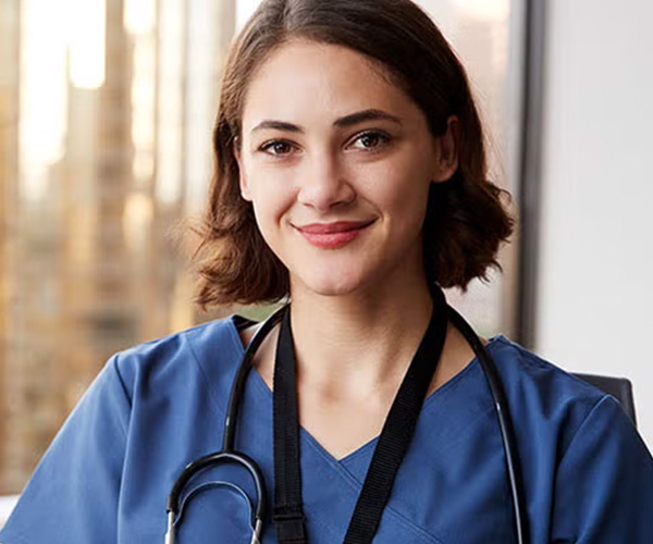 A female healthcare professional wearing blue scrubs and a stethoscope, seated in front of a window with a cityscape background.