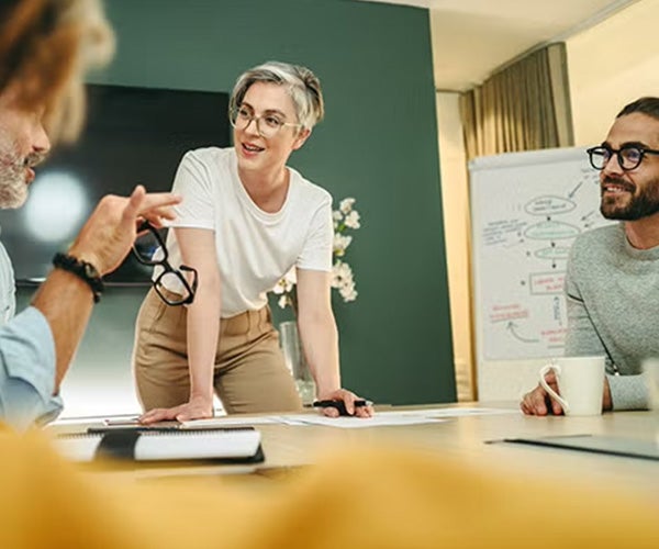 Group of people in a meeting, with a woman standing and engaging with others seated around a table.