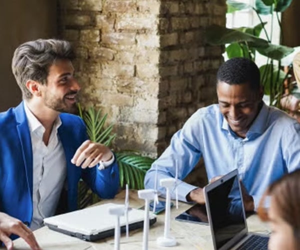 Two men laughing and talking while seated in a modern café-style workspace, with laptops and a relaxed atmosphere.