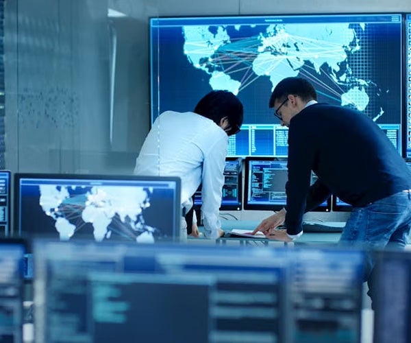 Two individuals in a control room reviewing data on multiple monitors, with world maps and data lines displayed.