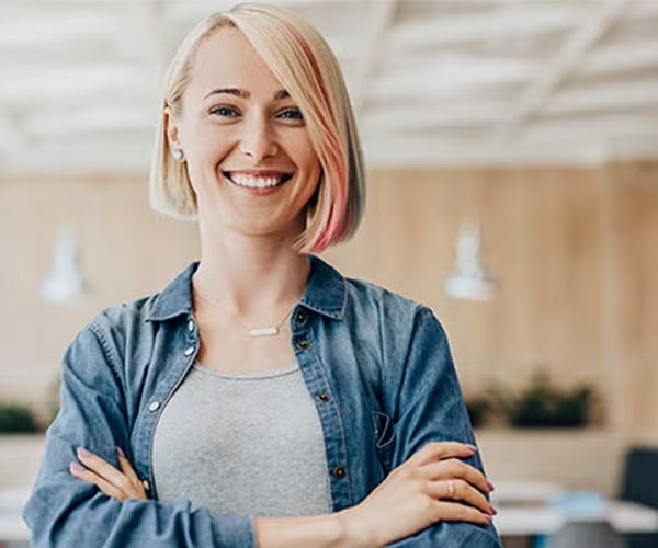 Smiling woman with blonde hair, some pink streaks, standing confidently with arms crossed in a bright office setting.