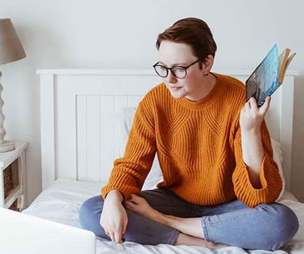 A student wearing glasses sitting on a bed, holding a book, with a laptop placed nearby.