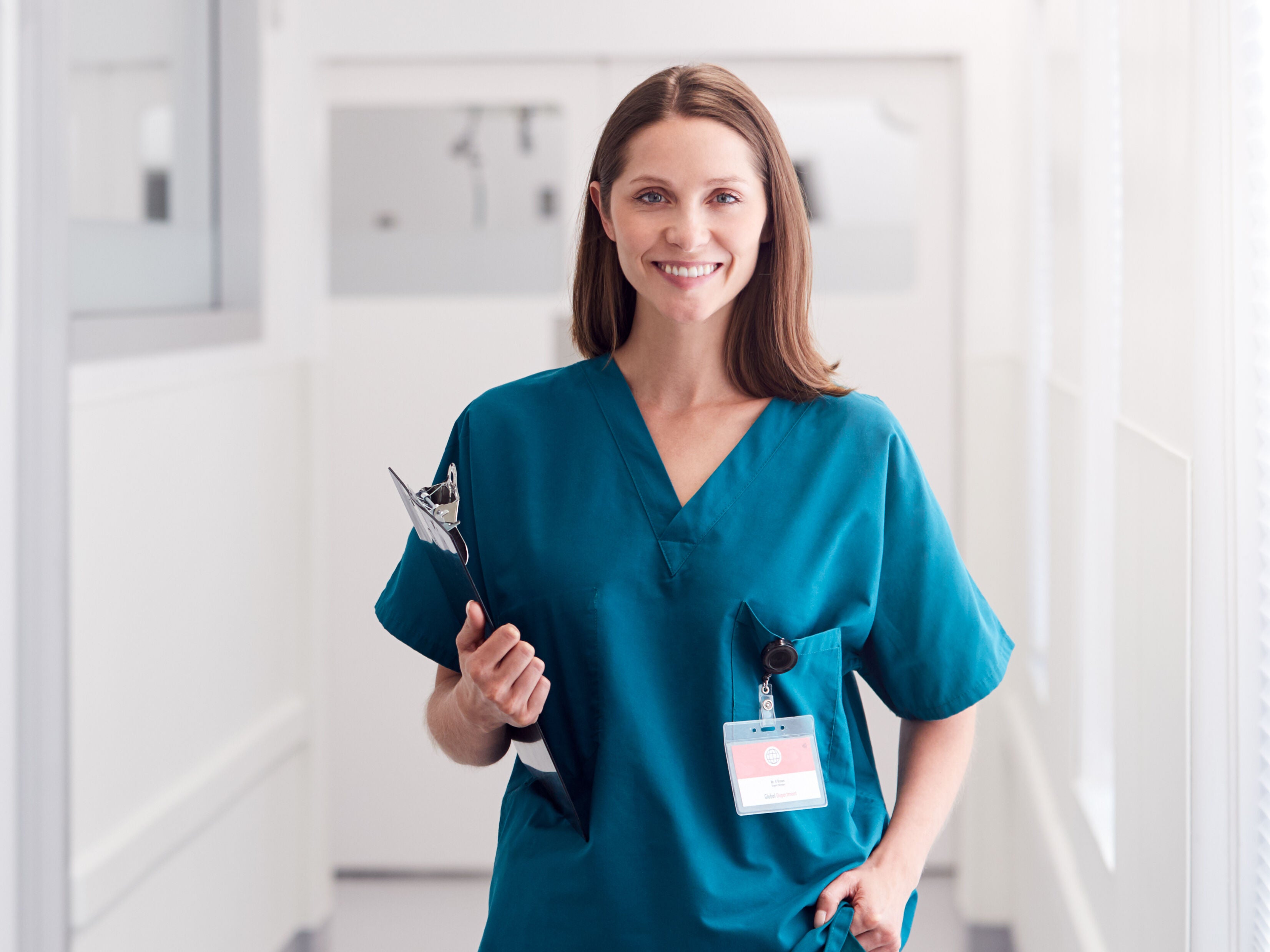 Portrait of smiling female doctor wearing scrubs standing in hospital corridor holding clipboard.