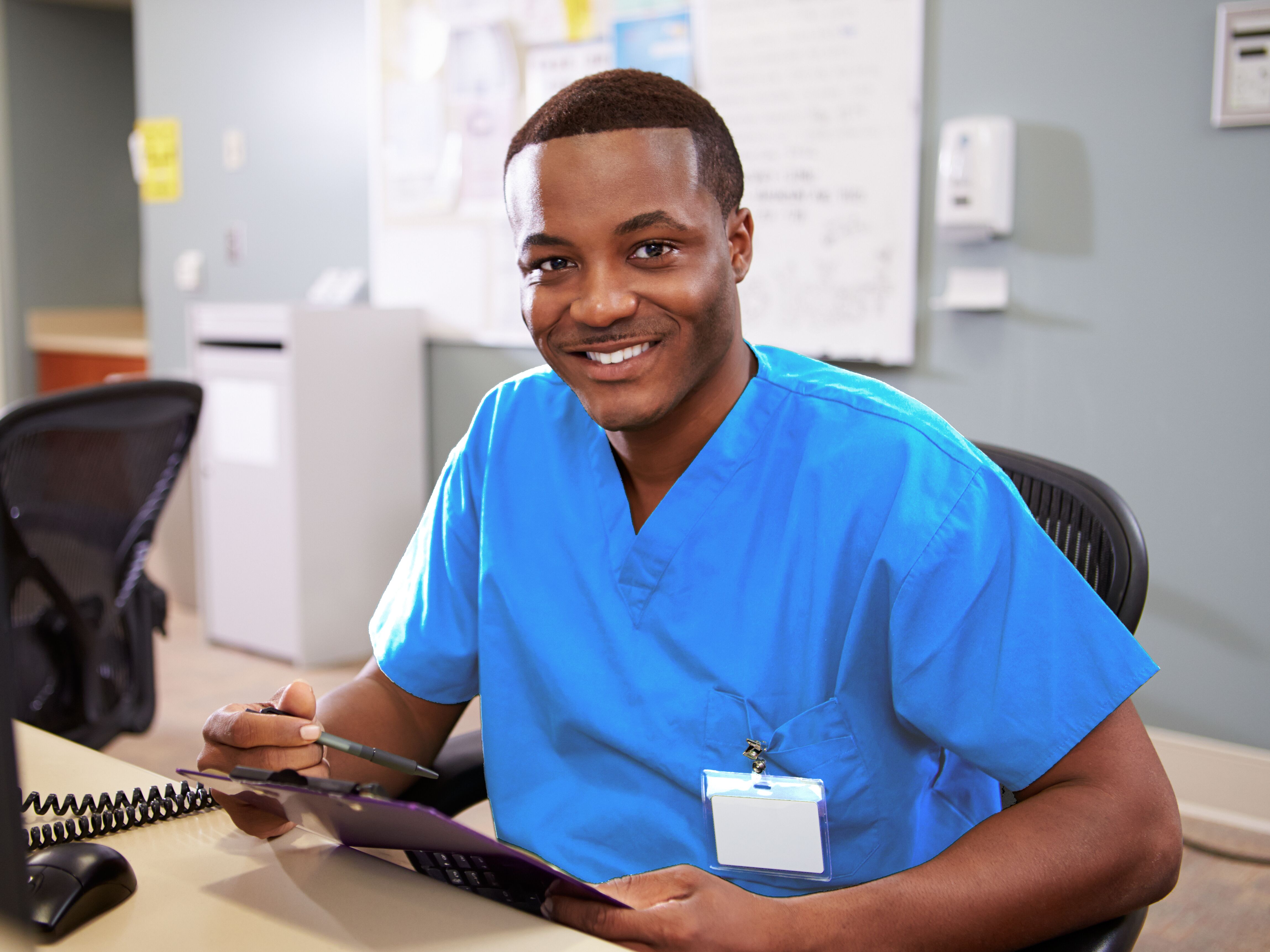 A cheerful male nurse in blue scrubs sitting at a hospital workstation, holding a pen and clipboard.
