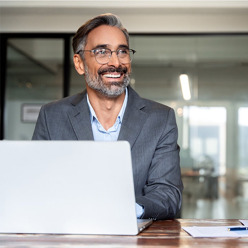 A senior man with gray hair and glasses sits at a wooden desk with a laptop, appearing engaged in work.