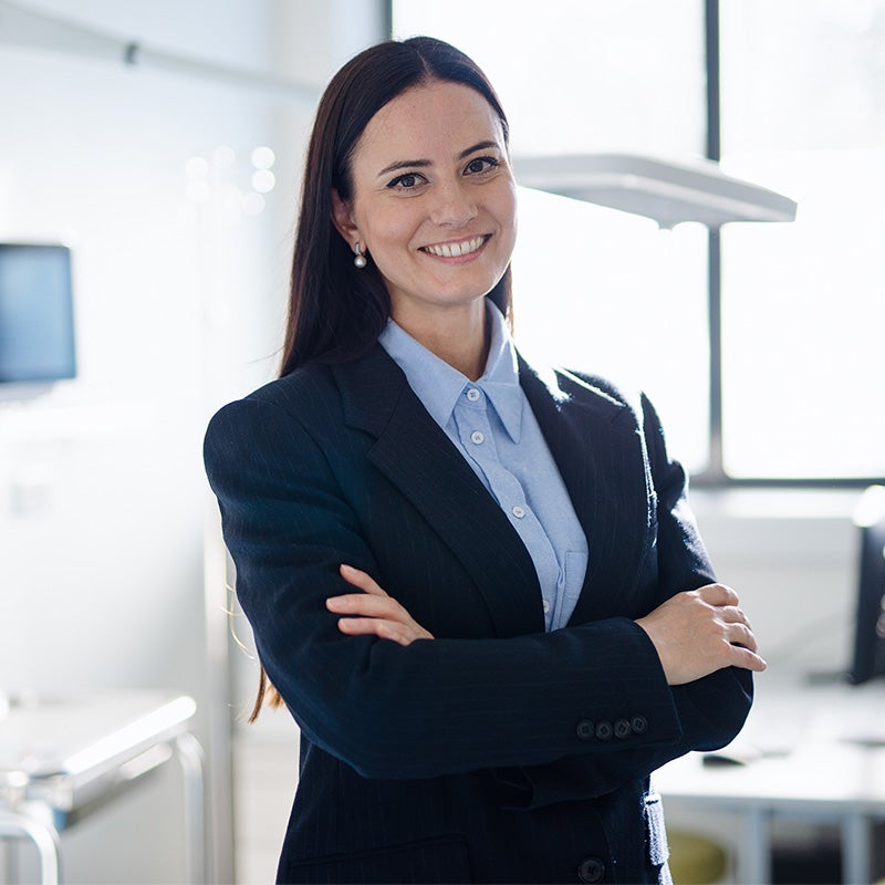 A professional woman in a dark business suit stands confidently with arms crossed in a bright office setting.