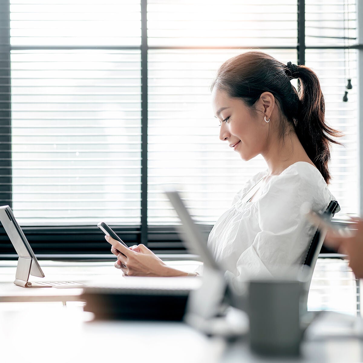 A young woman in a white blouse sitting at a desk, smiling while looking at her smartphone in a modern office setting.
